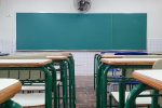 Vista de trás de uma sala de aula, com a lousa de frente para quem olha a imagem e um vão entre as carteiras, alinhadas lado a lado
